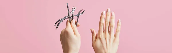 Vue recadrée d'une femme tenant un poussoir à cuticules et des pinces isolées sur une photo panoramique rose — Photo de stock