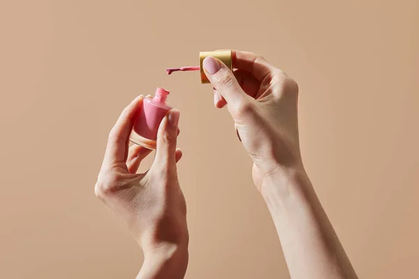 Vista parcial de la mujer sosteniendo botella abierta de esmalte de uñas rosa aislado en beige - foto de stock