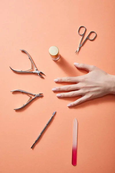 Vista recortada de la mano femenina con botella de esmalte de uñas e instrumentos de manicura en coral - foto de stock