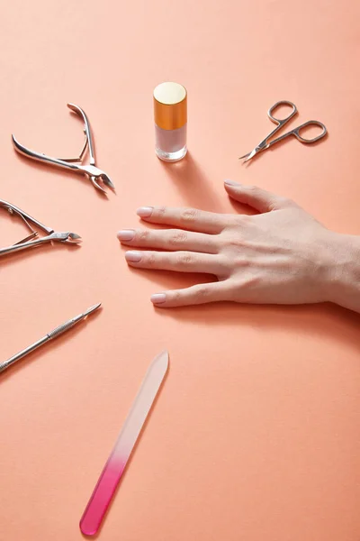 Vista parcial da mão feminina com garrafa de esmalte e instrumentos de manicure em coral — Fotografia de Stock