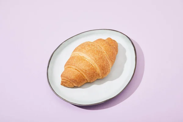 Croissant saboroso fresco na placa no fundo violeta — Fotografia de Stock