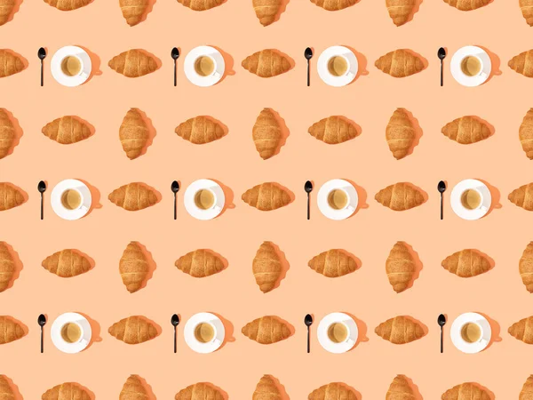 Von oben mit Löffeln, frischen Croissants auf Tellern und Kaffee auf orangefarbenem, nahtlosem Hintergrundmuster — Stockfoto