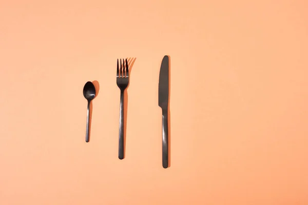 Вид сверху на металлическую блестящую черную вилку, ложку и нож на оранжевом фоне — стоковое фото