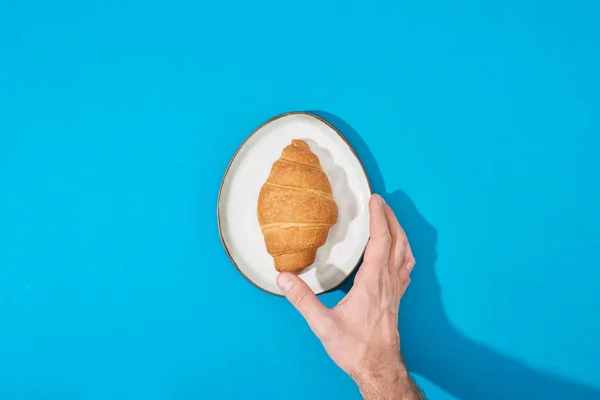 Cropped view of man holding fresh croissant on plate on blue background — Stock Photo