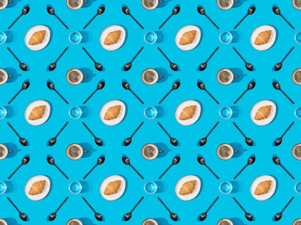 Vista superior de cucharas, croissants en platos, agua y café sobre fondo azul, sin costuras - foto de stock