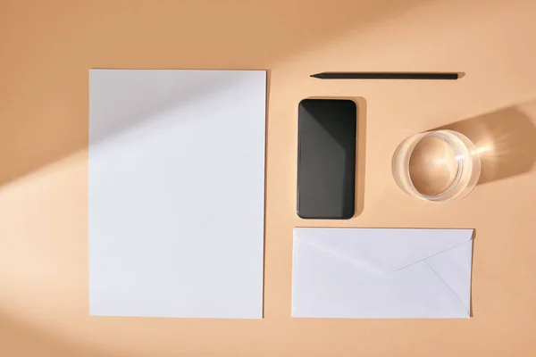 Posa piatta con foglio di carta, smartphone, matita, bicchiere d'acqua e busta su sfondo beige — Foto stock