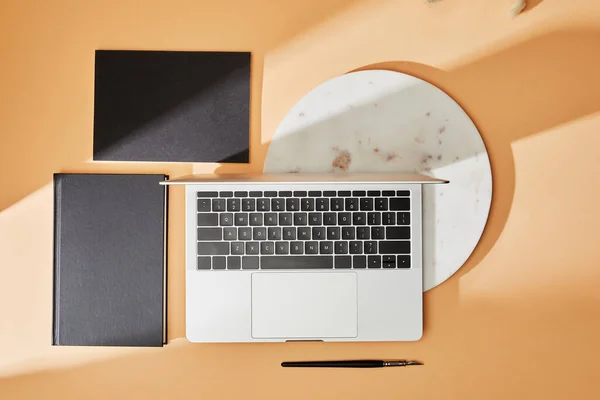 Top view of laptop, marble board, black notebooks and paintbrush on beige background — Stock Photo