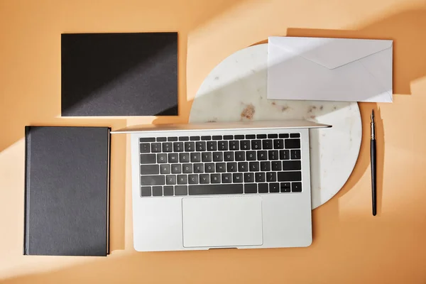 Вид зверху на ноутбук на мармуровій дошці, чорні блокноти, конверт і пензлик на бежевому фоні — стокове фото