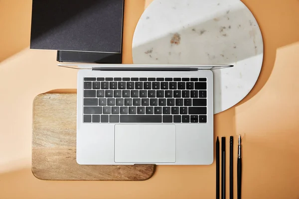 Vista superior de la computadora portátil en tablas de madera y mármol cerca de pinceles y cuadernos sobre fondo beige - foto de stock