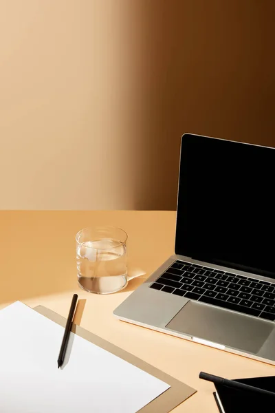 Computer portatile con schermo bianco vicino a vetro d'acqua, matita e carta su superficie beige — Foto stock