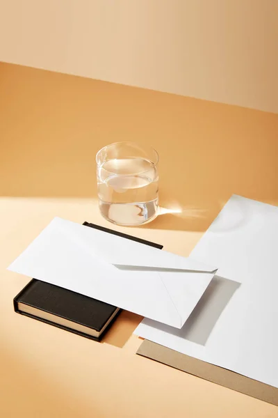 Vue grand angle de l'enveloppe sur carnet noir, feuilles de papier et verre d'eau sur fond beige — Photo de stock