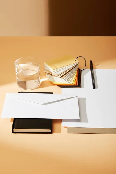 Envelope em caderno preto perto de folhas de papel, amostras de cores e vidro de água na superfície bege — Fotografia de Stock