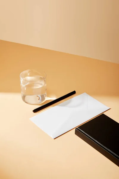 Vista de ángulo alto de sobre, pluma, vaso de agua y cuaderno negro en la superficie beige - foto de stock