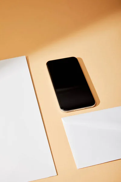 Vista de ángulo alto del teléfono inteligente cerca de sobre y hoja de papel sobre fondo beige - foto de stock
