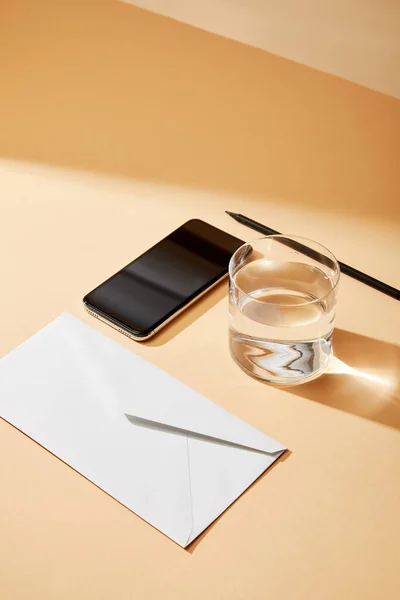 Hochwinkelaufnahme des Smartphones in der Nähe des Briefumschlags, Glas Wasser und Bleistift auf beigem Hintergrund — Stockfoto