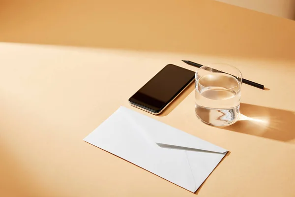 Високий кут зору смартфона, конверта, склянки води та олівця на бежевому фоні — стокове фото