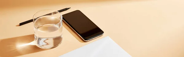 Panoramic shot of smartphone, envelope, glass of water and pencil on beige background — Stock Photo