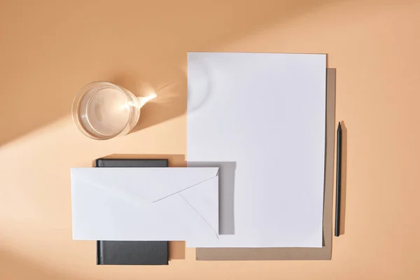 Vista superior de hojas de papel, sobre, pluma, vaso de agua y cuaderno sobre fondo beige - foto de stock