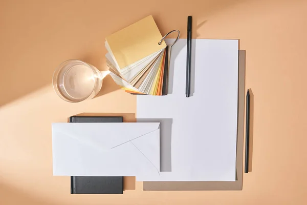 Flat lay com vidro de água, amostras de cores, folhas de papel, canetas, envelope e notebook sobre fundo bege — Fotografia de Stock