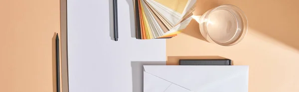 Tiro panorâmico de vidro de água, amostras de cores, folhas de papel, canetas, envelope e notebook sobre fundo bege — Fotografia de Stock