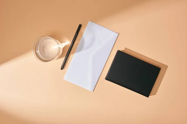 Vista dall'alto di busta, penna, bicchiere d'acqua e taccuino nero su sfondo beige — Foto stock