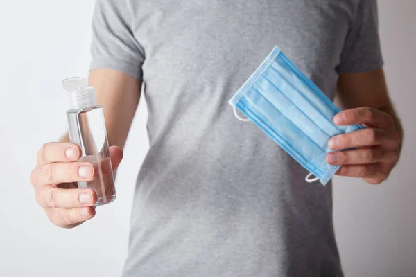 Visão recortada do homem segurando gel higienizador de mão em garrafa e máscara médica em fundo cinza — Fotografia de Stock