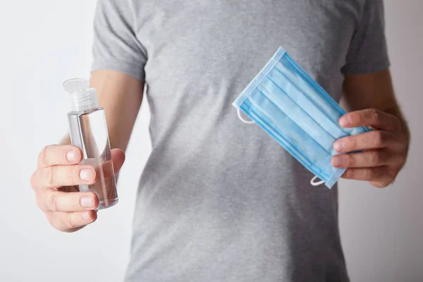 Cropped view of man holding gel hand sanitizer in bottle and medical mask on grey background — Stock Photo