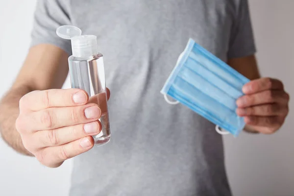 Vista recortada del hombre sosteniendo gel desinfectante de manos en botella y máscara médica sobre fondo gris - foto de stock