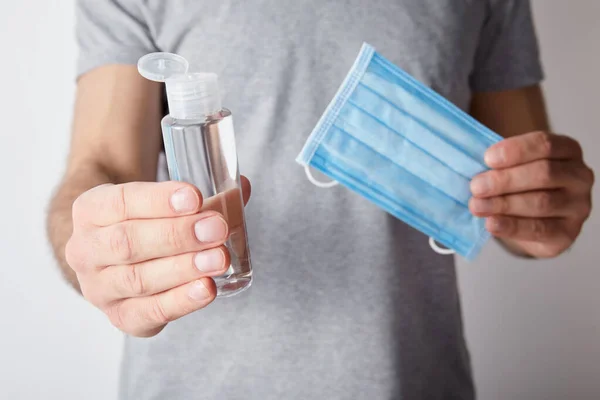Cropped view of man holding gel hand sanitizer in bottle and medical mask on grey background — Stock Photo