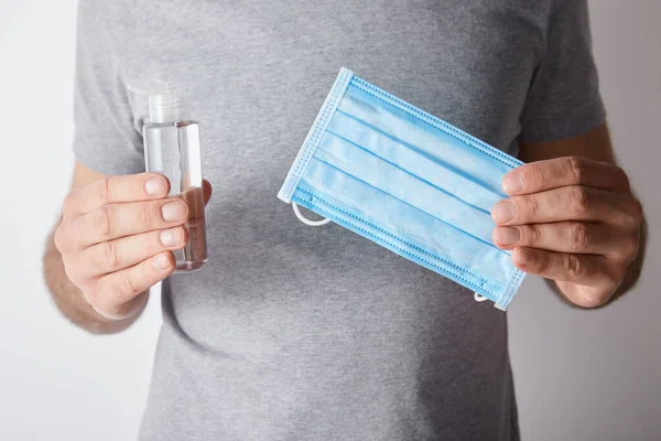 Vista recortada del hombre sosteniendo gel desinfectante de manos en botella y máscara médica sobre fondo gris - foto de stock