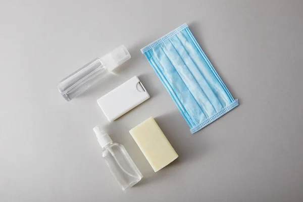 Vista superior del desinfectante de manos en botellas de spray, máscara médica y jabón antibacteriano sobre fondo gris - foto de stock