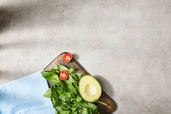Top view of plaid fabric near cutting board with basil leaves, cherry tomato and avocado halves on grey — Stock Photo