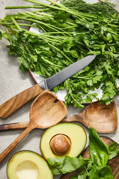 Вид сверху зелени на тарелку, нож, шпатели, половинки авокадо и листья авокадо на сером фоне — стоковое фото