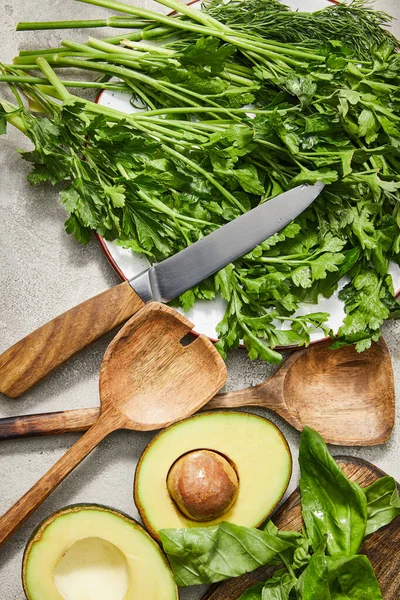 Vista superior de vegetación en placa, cuchillo, espátulas, mitades de aguacate y hojas de albahaca sobre fondo gris - foto de stock