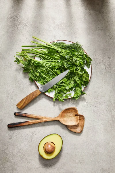 Вид сверху зелени на тарелке, нож, шпатели и половинки авокадо на сером фоне — стоковое фото