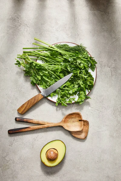 Vista dall'alto del verde su piatto, coltello, spatole e metà avocado su sfondo grigio — Foto stock