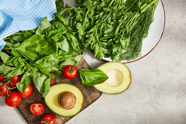 Vista superior de la tela cerca de la placa con vegetación y tabla de cortar con tomates cherry, hojas de albahaca y mitades de aguacate en gris - foto de stock