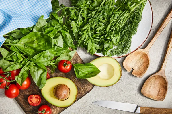 Vista superior de vegetação, folhas de manjericão, tomates cereja e metades de abacate perto de espátulas, faca e pano em fundo cinza — Fotografia de Stock