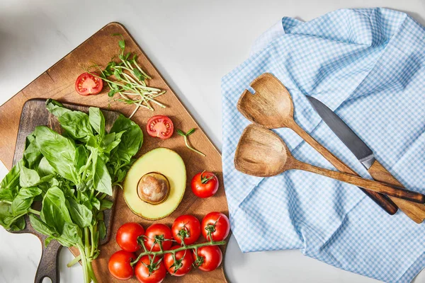 Vista superior de albahaca, tomates cherry, mitad aguacate y microgreens en tablas de cortar cerca de tela con espátulas y cuchillo en blanco - foto de stock