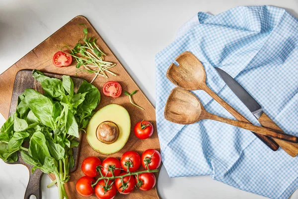 Vista superior de manjericão, tomate cereja, metade de abacate e microgreens em tábuas de corte perto de pano com espátulas e faca no branco — Fotografia de Stock