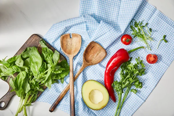 Vista superior de espátulas, perejil, microgreens, verduras y aguacate medio en tela a cuadros cerca de tabla de cortar con albahaca en blanco - foto de stock