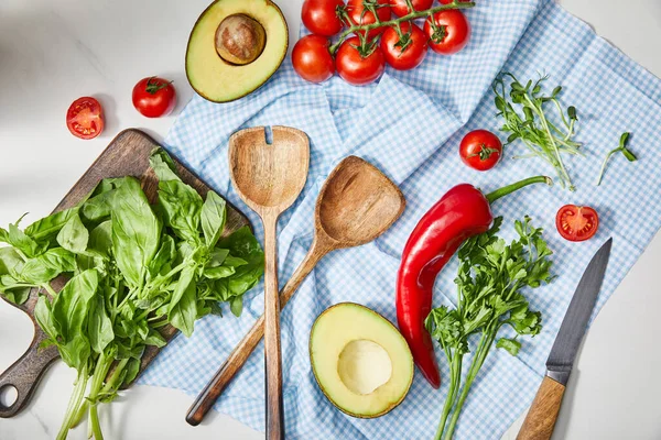 Vista superior de verduras, vegetación, mitades de aguacate, cuchillo y espátulas en tela cerca de albahaca en tabla de cortar en blanco - foto de stock