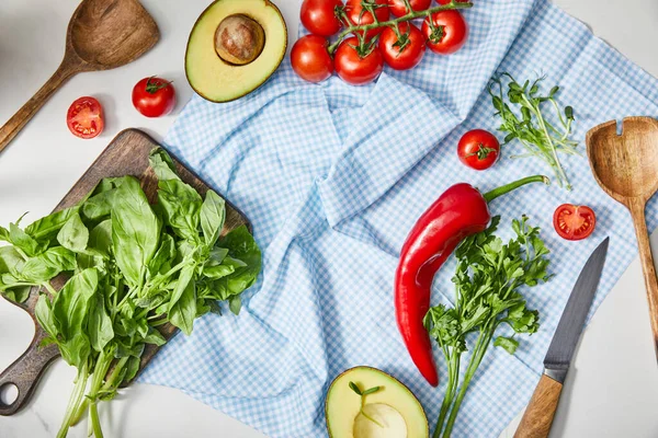 Vista superior de verduras, vegetación, mitades de aguacate, cuchillo y espátulas en tela a cuadros cerca de albahaca en tabla de cortar en blanco - foto de stock