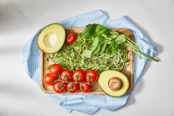 Draufsicht auf Schneidebrett mit Mikrogemüse, Basilikum, Kirschtomaten und Avocadohälften auf kariertem Tuch auf weiß — Stockfoto