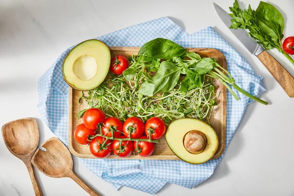 Vista superior da placa de corte com microgreens, manjericão, tomate cereja e abacate em pano perto de faca e espátulas em branco — Fotografia de Stock