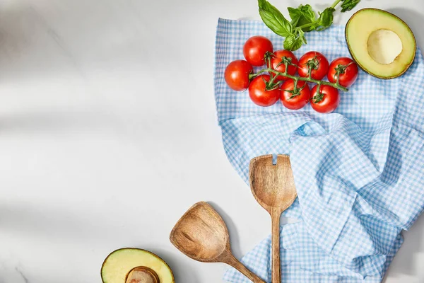 Vista superior de albahaca, tomates cherry, espátulas y mitades de aguacate con tela a cuadros sobre fondo blanco - foto de stock