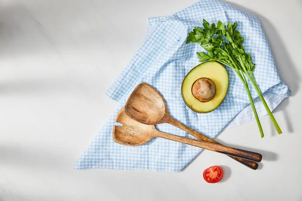 Top view of parsley and spatulas near avocado and cherry tomato halves with plaid cloth on white background — Stock Photo