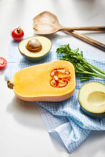 Vista en ángulo alto de verduras, vegetación y mitades de aguacate sobre tela cerca de espátulas sobre fondo blanco — Stock Photo