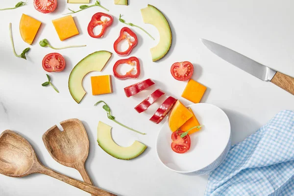 Vue du dessus du bol avec microgreens, légumes coupés et tranches d'avocat près du couteau, spatules et tissu sur blanc — Photo de stock