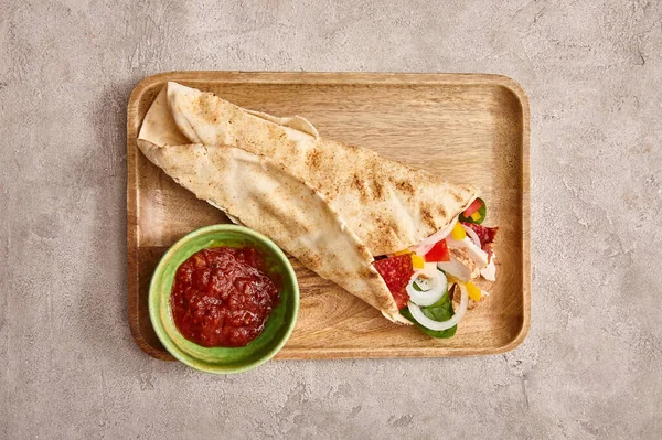 Vue du dessus du burrito frais avec poulet et légumes à bord près de la sauce chili sur fond gris béton — Photo de stock